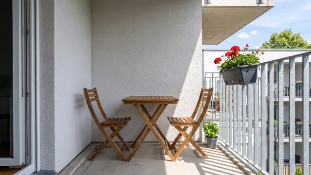 Symbolbild (KI-generiert): schmaler Balkon mit verzinktem Geländer, klappbarem Holztisch und zwei Klappstühlen, Balkonkasten mit Geranien, Sommersonne