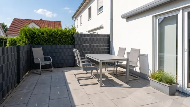 Symbolbild (KI-generiert): Terrasse mit Esstisch und Stühlen zwischen Hauswand mit Terrassentür und Sichtschutzzaun mit Thuja, Sommersonne
